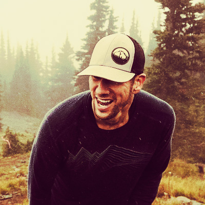 A man in a khaki baseball hat and dark shirt hunches over smiling as he climbs a steep hill in front of evergreen trees.